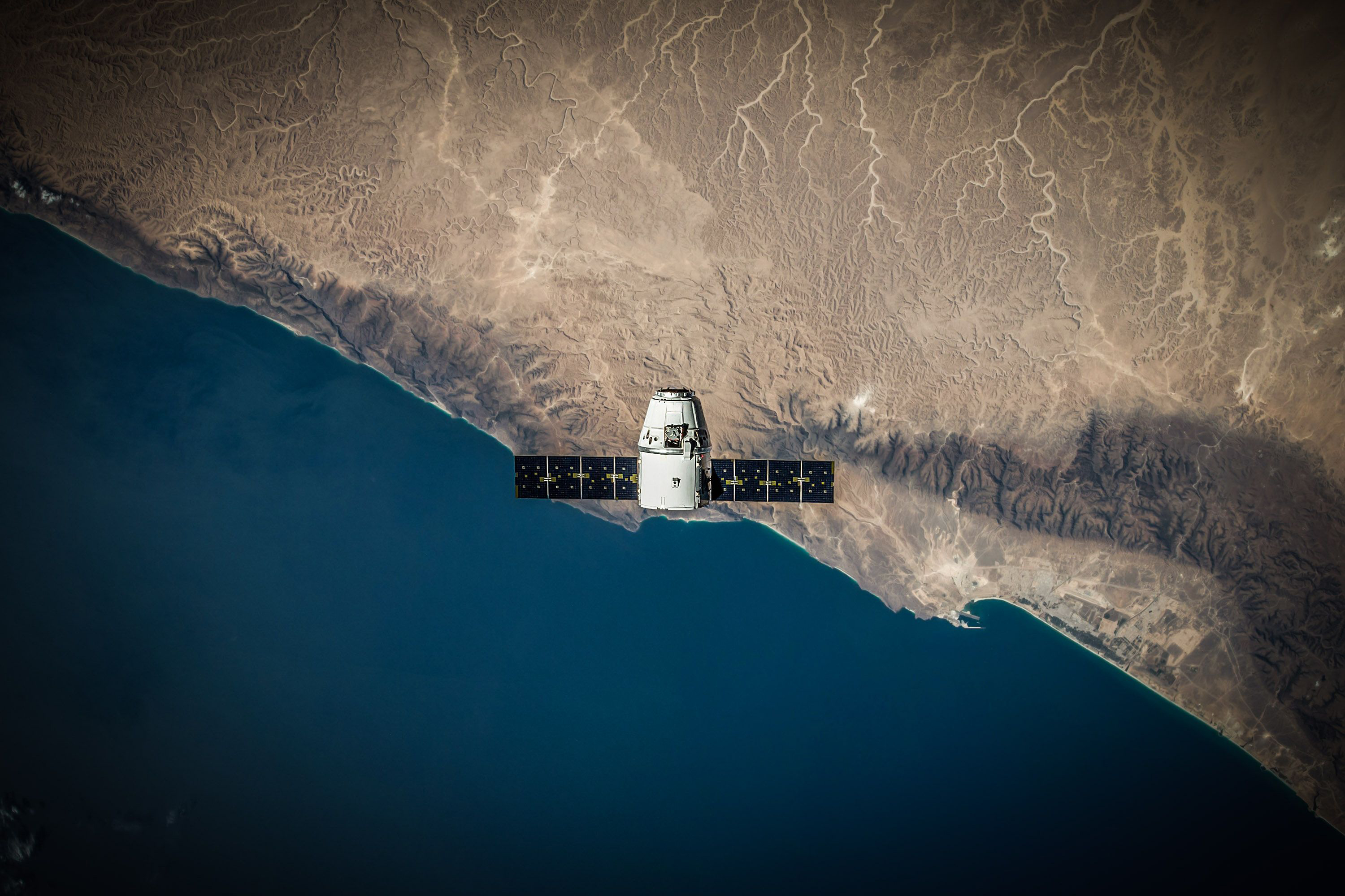 Cargo spacecraft with solar arrays orbiting above a coastline